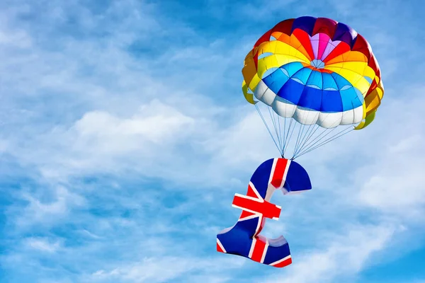 Pound of sterling symbol of British currency lifting by parachute on background of blue sky.