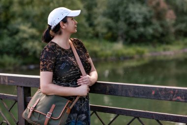 Omuz çantalı ve beyaz şapkalı bir kadın yazın parkta yürüyor..