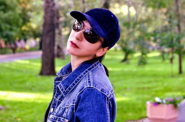 Yaz parkında kot ceket, pembe beyzbol şapkası ve güneş gözlüğü takan bir kadın..