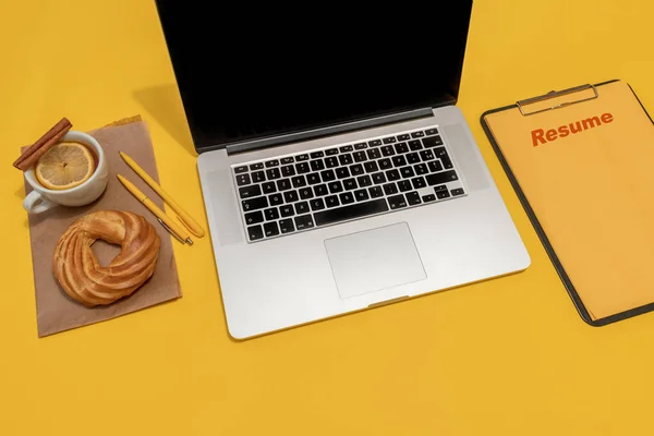 Laptop, cake, cup of tea and resume sheet on the Fortuna Gold Yellow Colour Background, top view.