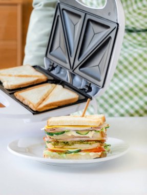 Eller tost makinesini, kızarmış taze ekmek dilimleriyle birlikte beyaz tabakta hazır sandviçin yanında tutuyor.