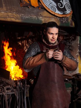 Viking demircide silah ve kılıç dövüyor. Savaşçı kıyafetli bir adam ateşin arka planında demircide..