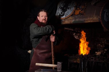 Viking demircide silah ve kılıç dövüyor. Savaşçı kıyafetli bir adam ateşin arka planında demircide..