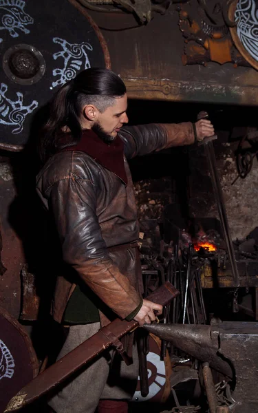 Viking demircide silah ve kılıç dövüyor. Savaşçı kıyafetli bir adam ateşin arka planında demircide..