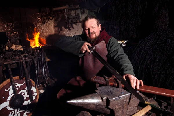 Viking forges weapons and swords in the smithy. A man in a warrior\'s clothes is in the smithy on the background of fire.