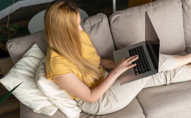 Orta yaşlı sarışın kadın kanepede oturuyor ve internetten bilgisayarla iletişim kuruyor..