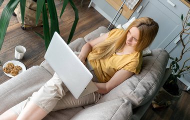 Orta yaşlı sarışın kadın kanepede oturuyor ve internetten bilgisayarla iletişim kuruyor..