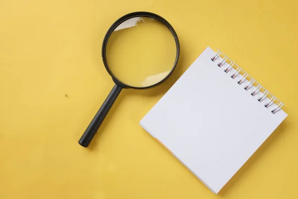 flat magnifying glass and white spiral notepad on a table yellow background