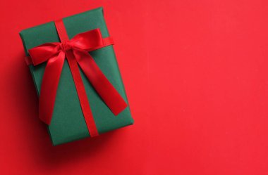 Kırmızı arkaplanda kırmızı kurdeleli yeşil bir hediye kutusu.