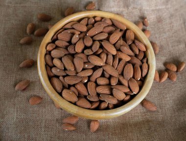 Üst manzara, beyaz arkaplanda izole edilmiş keten kumaş üzerinde ahşap bir kasede duran kahverengi ham badem yığını.