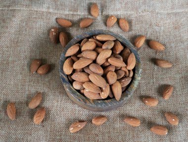 Beyaz arka planda izole edilmiş keten kumaş üzerinde ahşap bir kasede duran kahverengi çiğ badem yığını.