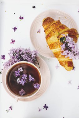 Kahvaltı havasında. Pembe kahve ve leylak çiçekli Fransız kruvasanı. Bahar geldi. Üst Manzara. Seçici odak sanat gürültüsü 