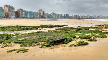 Arka planda binalar olan boş bir sahilde kum ve kayalar. Gijn, Asturias, İspanya