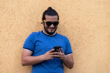 Güneş gözlüklü genç Latin adam telefonda mesaj atıyor ve gülümsüyor. Teknoloji konsepti