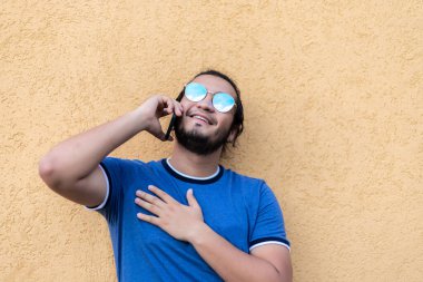 young bearded latin guy with sunglasses leaning against a yellow wall talking on the phone. Fresh air