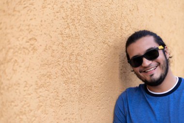 young latin guy with glasses and long hair, smiling looking at the camera, leaning against a yellow wall. Latin life