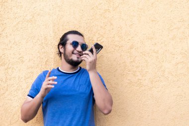 Genç, sakallı, güneş gözlüklü, sarı duvara yaslanmış, sesli mesaj yollayan Latin bir adam. Temiz hava.