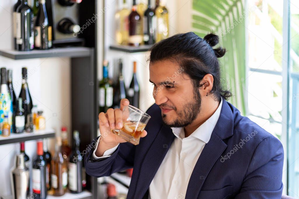 young latino man sitting on home cabinet, drinks whiskey. New normal.