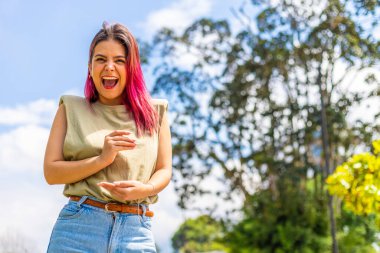 Genç Latin bir kadın kameraya bakıyor, halka açık bir parkta duruyor ve el hareketi yapıyor..