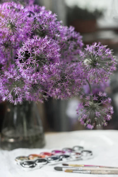 Bouquet d'oignons décoratifs fleuris de couleur violet-lilas dans un vase en verre se tient sur la table à côté des pinceaux et des peintures à l'aquarelle — Photo