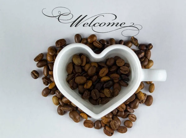 Etrafında kahve çekirdekleri olan beyaz kalp şeklinde bir kupa ve yazıtlara hoş geldin..