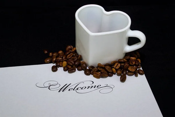 Etrafında kahve çekirdekleri ve hoş geldin işareti olan kalp şeklinde beyaz bir kupa..