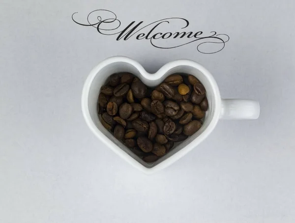 İçinde kahve çekirdekleri olan kalp şeklinde bir kupa beyaz bir çarşafın üzerinde karşılama yazısıyla..