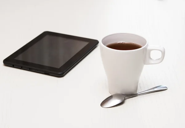 White cup of tea, digital tablet and spoon on light wooden desk. — Stock Photo, Image