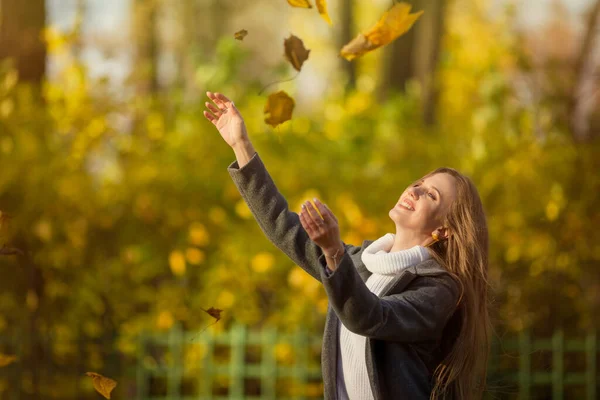 Beautiful happy woman with light brown hair smiles and raises her hands. Leaf fall in the autumn park. Attractive young woman in a gray coat and white sweater. Yellow foliage, Fall season. Autumn mood.