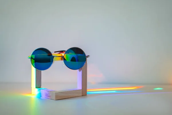 Blue reflective sunglasses on wooden stand in front of a white background with rainbow reflections on the lenses and on the stand. Front view