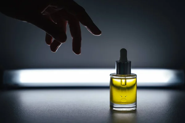 Backlit yellow face oil with a dropper bottle on a grey background with a reaching female hand. Side view