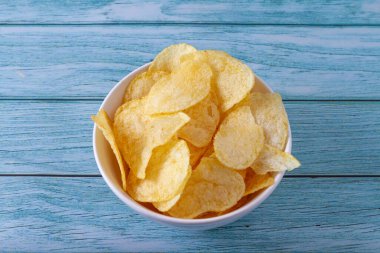 Atıştırmalık konsepti. Beyaz bir kasede patates cipsi, ahşap bir masada mavi tonda..