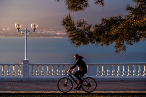 男は柵や提灯で堤防に沿って自転車に乗る。夕日があなたの顔に輝いています。背景に海と山 — ストック写真