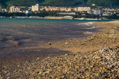 Tatil köyünün çakıl taşlı plajında bir dalga akıyor. Deniz köpüğü, çakıl taşı. Arka planda, bulutlarla kaplı dağlar. Dağların eteğindeki şehir