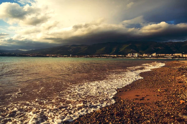 Er loopt een golf over het kiezelstrand van de badplaats. Zeeschuim, kiezels. Op de achtergrond, bergen bedekt met wolken. Stad aan de voet van de bergen — Stockfoto