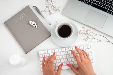 Ofis dairesi: bilgisayar, modern klavye, not defteri, kablosuz kulaklıklar, şık mumlar ve beyaz bir ofis masasında kurumuş çiçekler. Kırmızı manikürlü kız klavyeye yazı yazıyor..