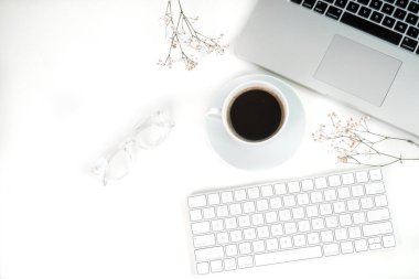 Ofis dairesi: bilgisayar, modern klavye, gözlükler, bir fincan kahve ve beyaz ofis masasında kurumuş çiçekler.