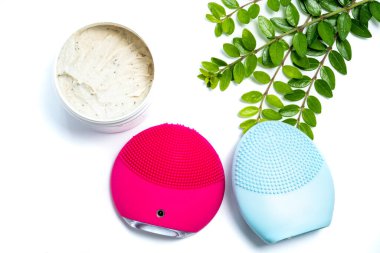 Pembe ve mavi yüz temizleme fırçası, yüz fırçası ve beyaz arka planda yeşil yapraklar.