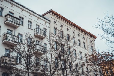 Berlin, Almanya 'da bulutlu gökyüzünün altında ön planda çıplak ağaçlarla modern beyaz apartman manzarası düşük açılı.