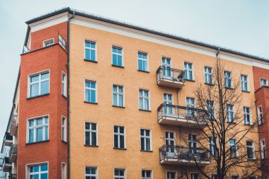Ön planda balkonu ve çıplak ağacı olan birden fazla katlı apartmanın köşesindeki kutucuk tipi katlama pencereleri