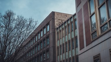 Almanya 'da bulutlu gökyüzünün altında önü çıplak ağaçla kaplı iki uzun ofis binası arasında dar pencereli bir daire.
