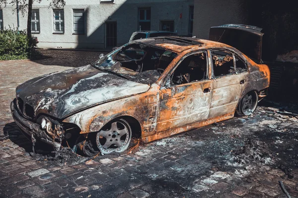 Yanmış, tamamen harap olmuş lüks bir sedan. Yanmış bir apartman bloğunun asfaltlı avlusunda, önü manzaralı.