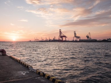 Gün batımında, renkli gökyüzüne karşı kargo siluetini yüklemek için devasa endüstriyel vinçlerle bir tersanenin sularına bakın.
