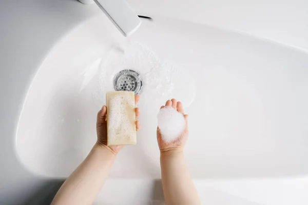 Çocuk ellerini suyla beyaz bir lavaboda sarı sabunla yıkıyor. Üst görünüm.