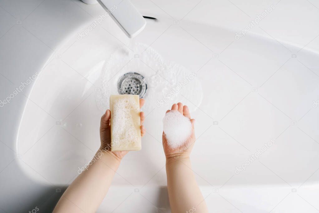 The child washes his hands with yellow soap in a white sink with water. Top view.