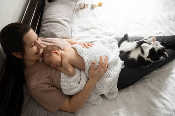 Harika zaman geçirdim. Genç anne yeni doğmuş bebeğiyle eğlenirken, yatakta uzanıp onu kucaklıyor. Kadının bacaklarında yatan sevimli evcil kedi. Mutlu çocukluk kavramı 