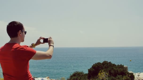 Jeune homme assis sur des rochers près de la mer et film vidéo avec téléphone portable — Video