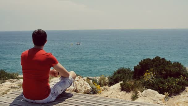 Jeune homme assis sur une falaise et regardant vers la mer — Video