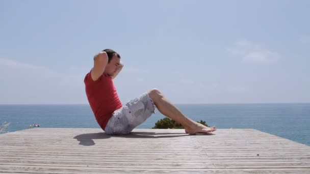 Atletische blanke man pompen de buikspieren op rotskust strand op zonnige dag — Stockvideo