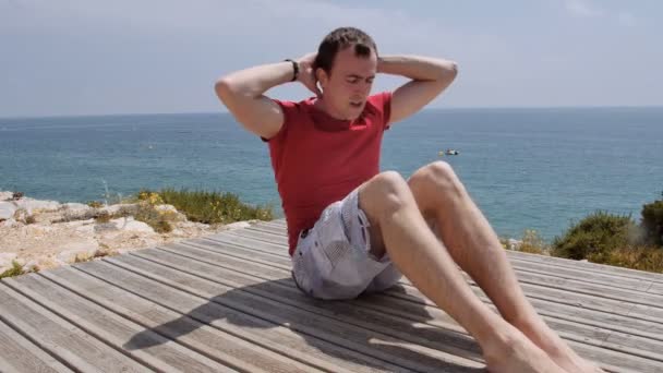 Athletischer kaukasischer Mann pumpt an sonnigen Tagen die Bauchmuskeln am felsigen Küstenstrand — Stockvideo